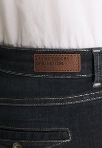 Tummansiniset farkut, joissa on nahkainen etiketti, jossa lukee "UNITED COLORS OF BENETTON". Ominaisuuksiin kuuluu näkyvät saumat ja nappidetaili vyötäröllä.