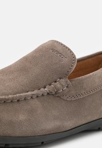 Zapatilla gris de ante de tipo slip-on con detalles de costura, que presenta un logo de Geox embellecido en el lateral y una suela de goma para mayor tracción.
