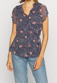 Blouse marine à fleurs avec manches courtes, encolure à volants et taille à basque. Présente des motifs floraux vibrants en rose et blanc sur un tissu transparent.