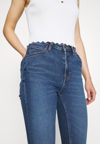 Jeans de mezclilla azul de talle alto con una textura desgastada, cinco bolsillos y un sutil detalle de costura, combinados con un top corto de canalé blanco.