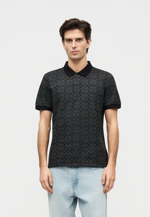 Mladý muž v čiernej polo košeli s krátkym rukávom s potlačou, čiernym golierom a svetlomodrými džínsami, stojaci pred jednofarebným pozadím.