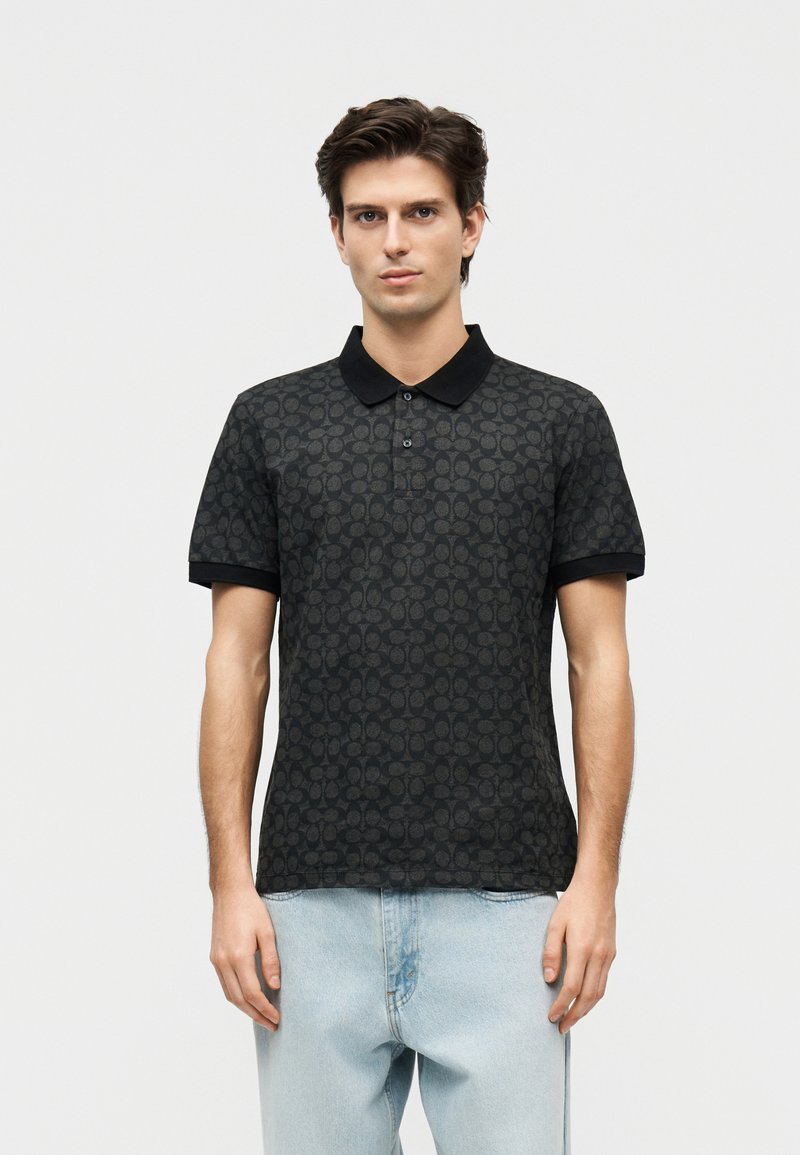 Jeune homme portant un polo à manches courtes noir à motifs avec un col noir et un jean bleu clair, debout devant un fond uni.