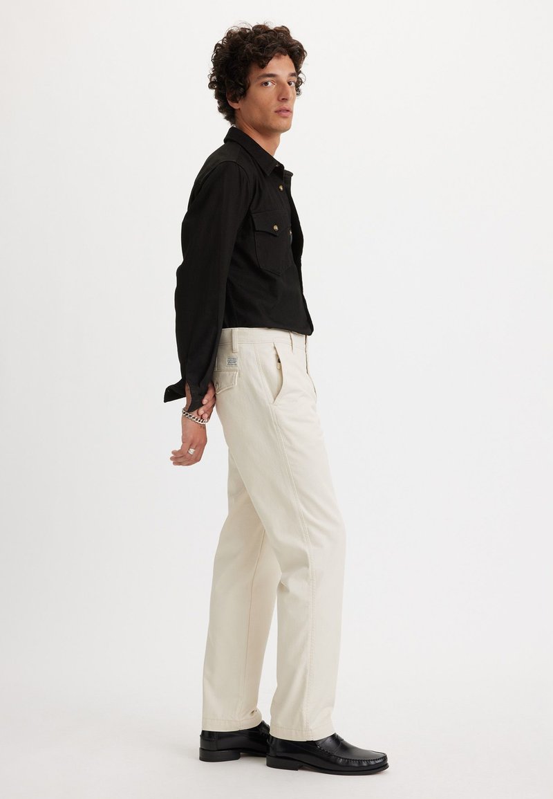 Homme en chemise noire boutonnée, pantalon crème et chaussures noires se tient de profil avec les mains derrière le dos sur fond blanc.