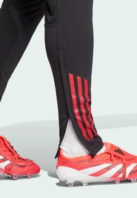 Black athletic pants with red vertical stripes, featuring side zippers. Paired with bright red soccer cleats with white accents.