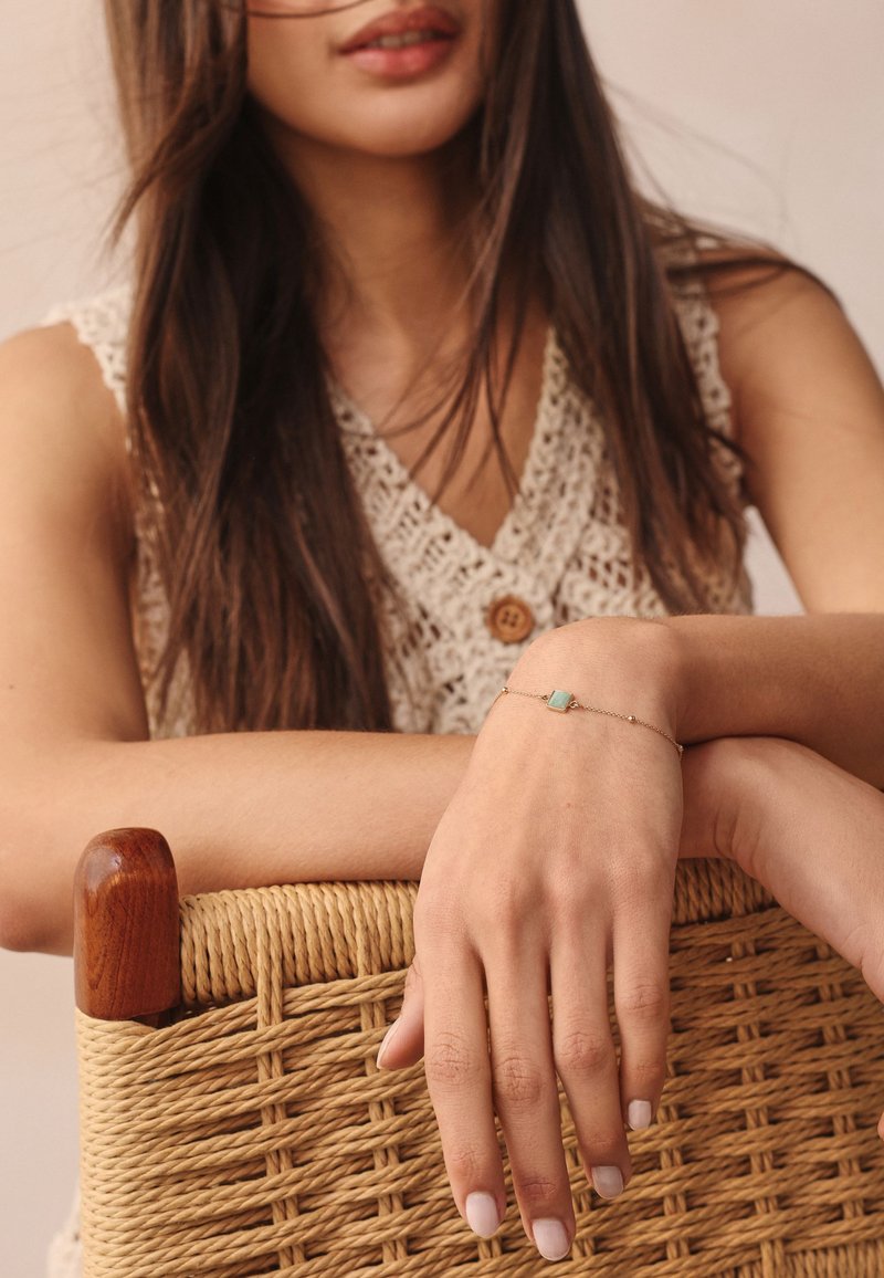 Gold chain bracelet featuring a small turquoise stone, worn on a delicate wrist. Background includes a woven, light-colored basket.