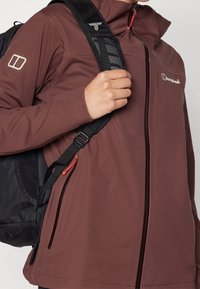 Personne portant une veste Berghaus marron tenant la sangle d'un sac à dos noir sur son épaule.