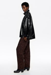 Čierna kožená oversize bunda, hnedé nohavice so širokými nohavicami a leopardím vzorom, a čierne podpätkové topánky; model je v profile na neutrálnom pozadí.