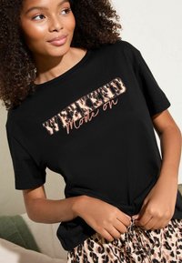 Zwarte T-shirt met "WEEKEND" in vette luipaardprint en cursief "Mode aan." Korte mouwen, relaxte pasvorm, gecombineerd met luipaardprint onderstukken.