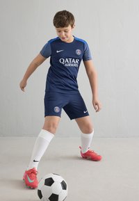 Fotbalový dres tmavě modré barvy se světle modrými doplňky, s logem "Qatar Airways". Hráč nosí jasně růžové kopačky a bílé ponožky a manipuluje s fotbalovým míčem.