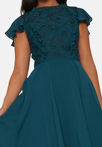 Tealfärgad klänning med en strukturerad överdel som har blommig spets och volangdetaljer, som flödar ner i en plisserad kjol med korta svävande ärmar.