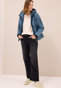 Giacca isolante blu con cappuccio, maglione beige, jeans neri e sneaker crema. Caratterizzata da tasche con zip e un design strutturato.
