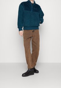 Mörk teal fleece tröja med kvartdragkedja och ribbad textur, kombinerad med bruna vida byxor och svarta ankelstövlar.