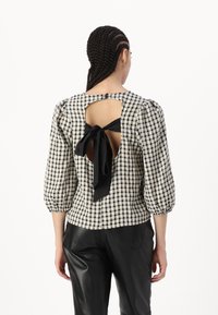 Blusa a quadri in bianco e nero con maniche a sbuffo, schiena scoperta e dettaglio con cravatta nera. Realizzata in tessuto leggero. Abbinata a pantaloni in pelle.