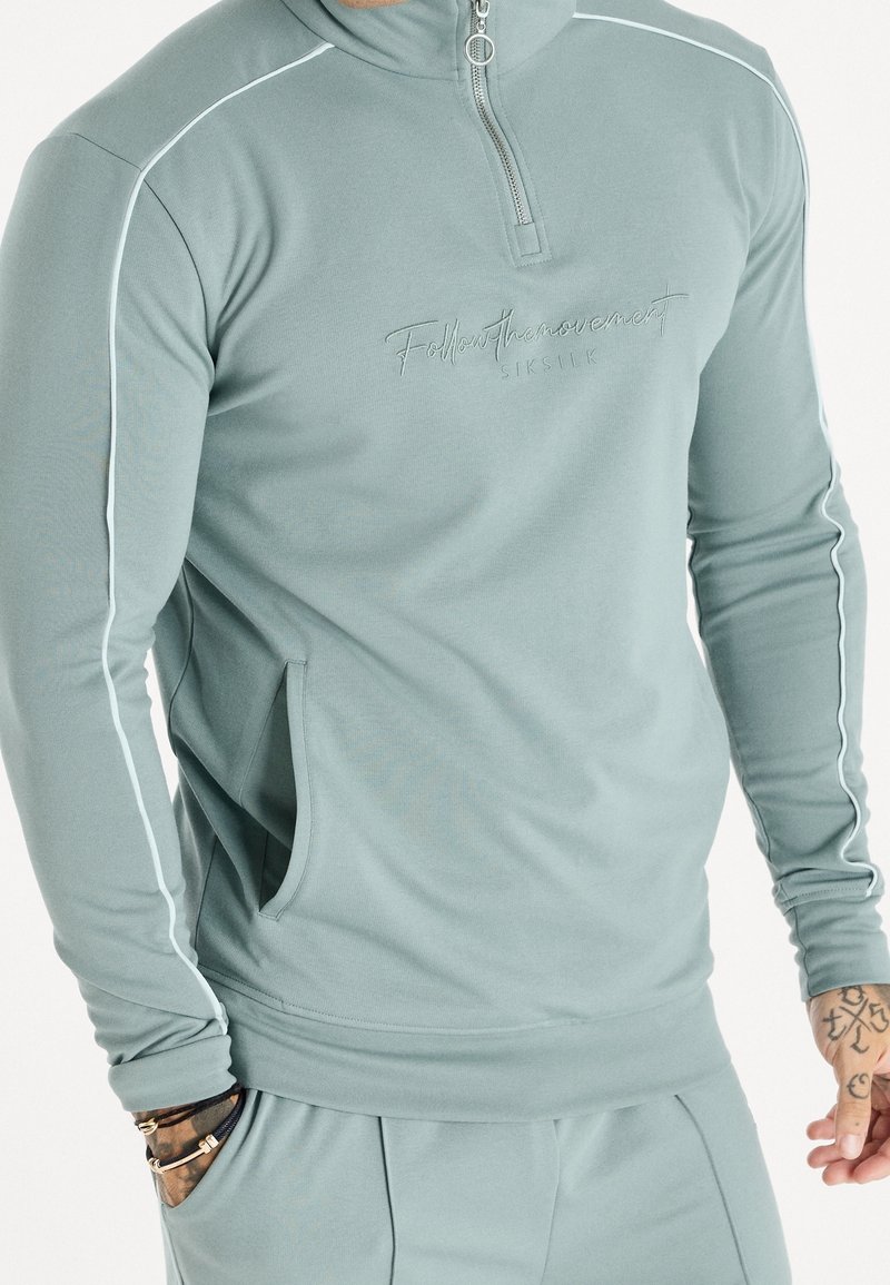 Sweat-shirt vert clair à fermeture éclair, fabriqué en tissu lisse, présentant un logo brodé discret et un liseré blanc le long des côtés, avec des poches latérales.