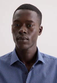 Giovane uomo con capelli corti che indossa una camicia blu a pois bianchi, rivolto in avanti con un'espressione neutra su uno sfondo semplice.