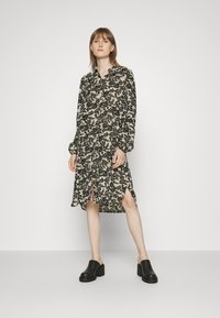 Vestido de manga longa em bege com padrão floral preto, apresentando colarinho com botões, fendas laterais e um ajuste solto. Combinado com mules de salto bloco pretas.