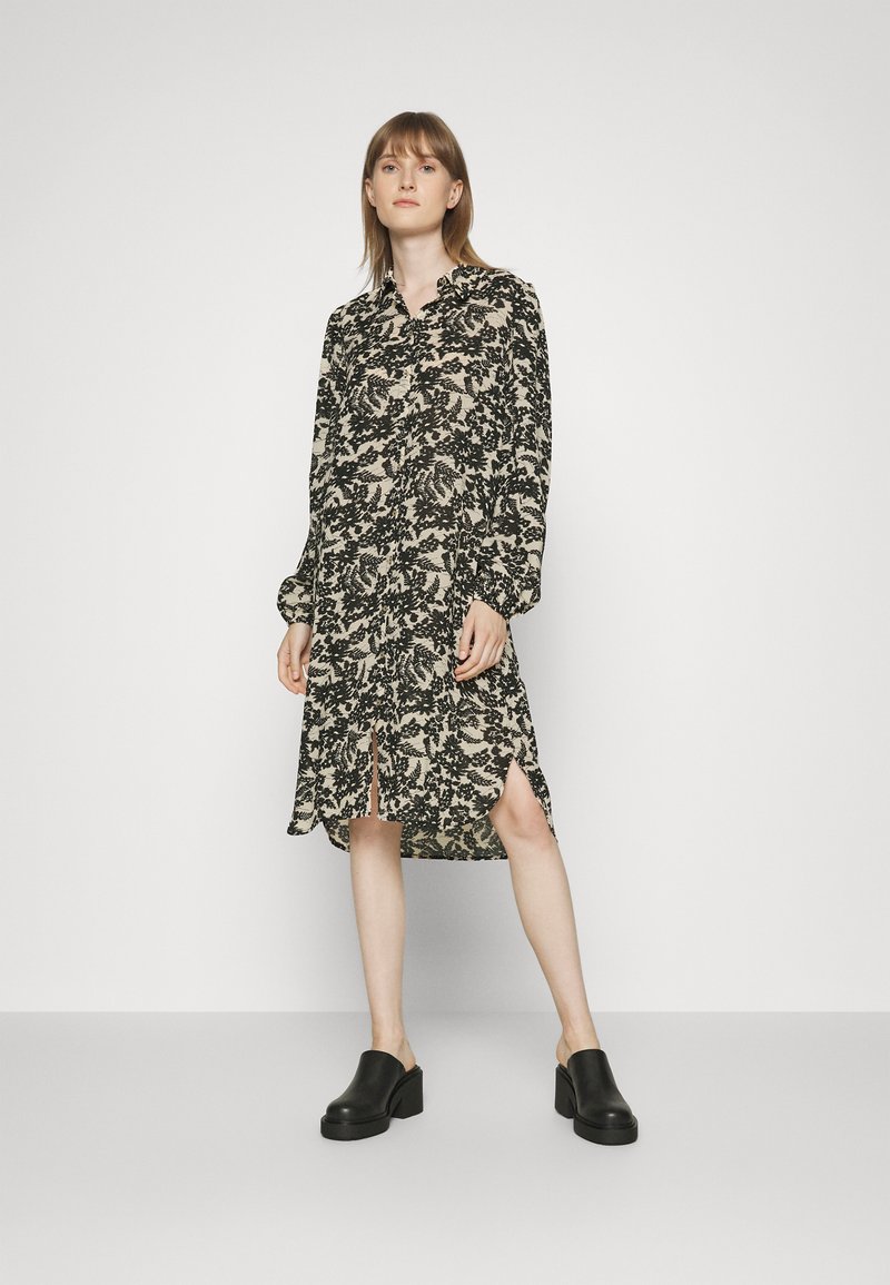 Vestido de manga longa em bege com padrão floral preto, apresentando colarinho com botões, fendas laterais e um ajuste solto. Combinado com mules de salto bloco pretas.