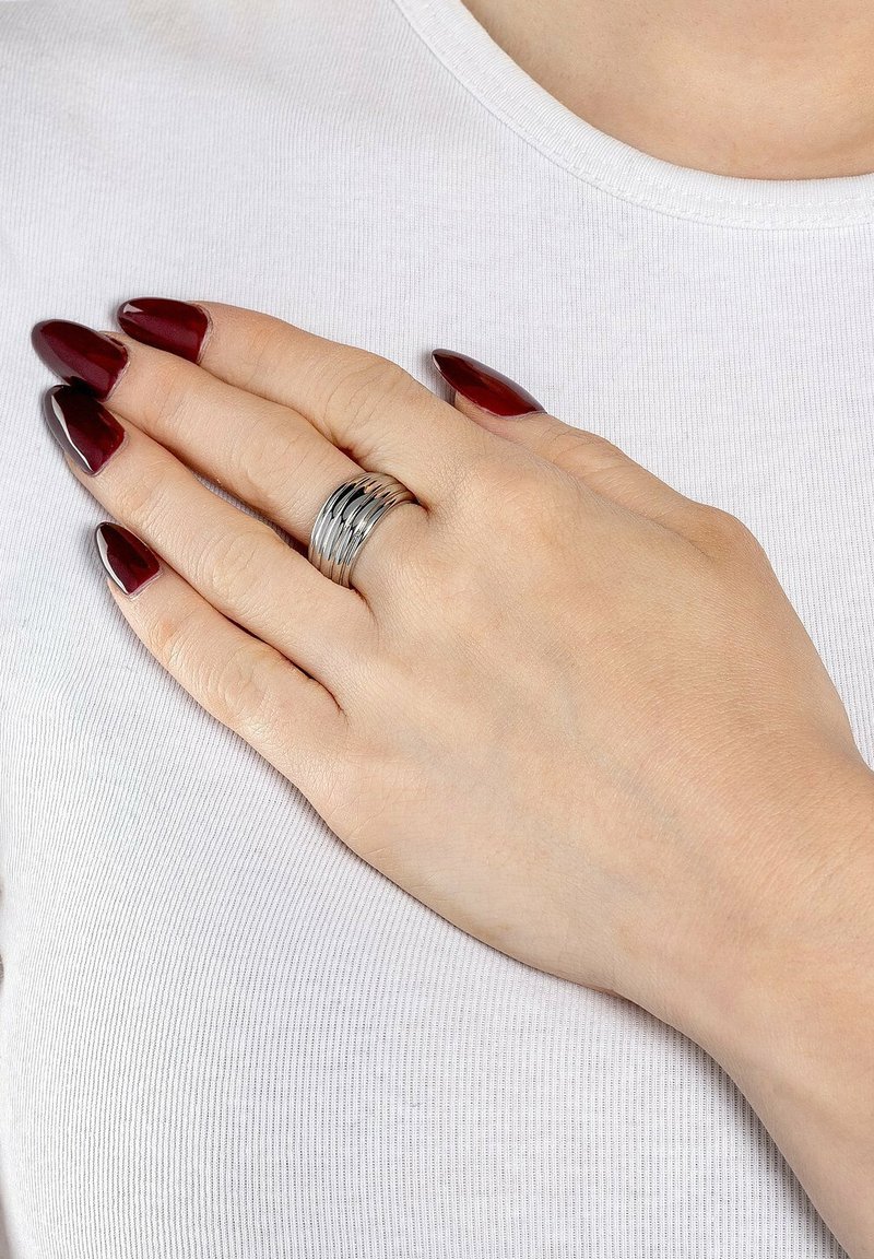 Sølv multi-band ring med stablet design, der reflekterer lys, båret på en hånd med lange, burgunder manikurerede negle mod en hvid top.