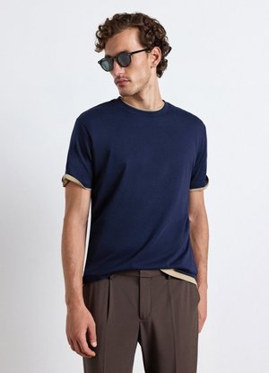 Uomo con capelli ricci che indossa occhiali da sole neri, una maglietta blu navy con maniche risvoltate e pantaloni marroni, in piedi davanti a uno sfondo semplice.