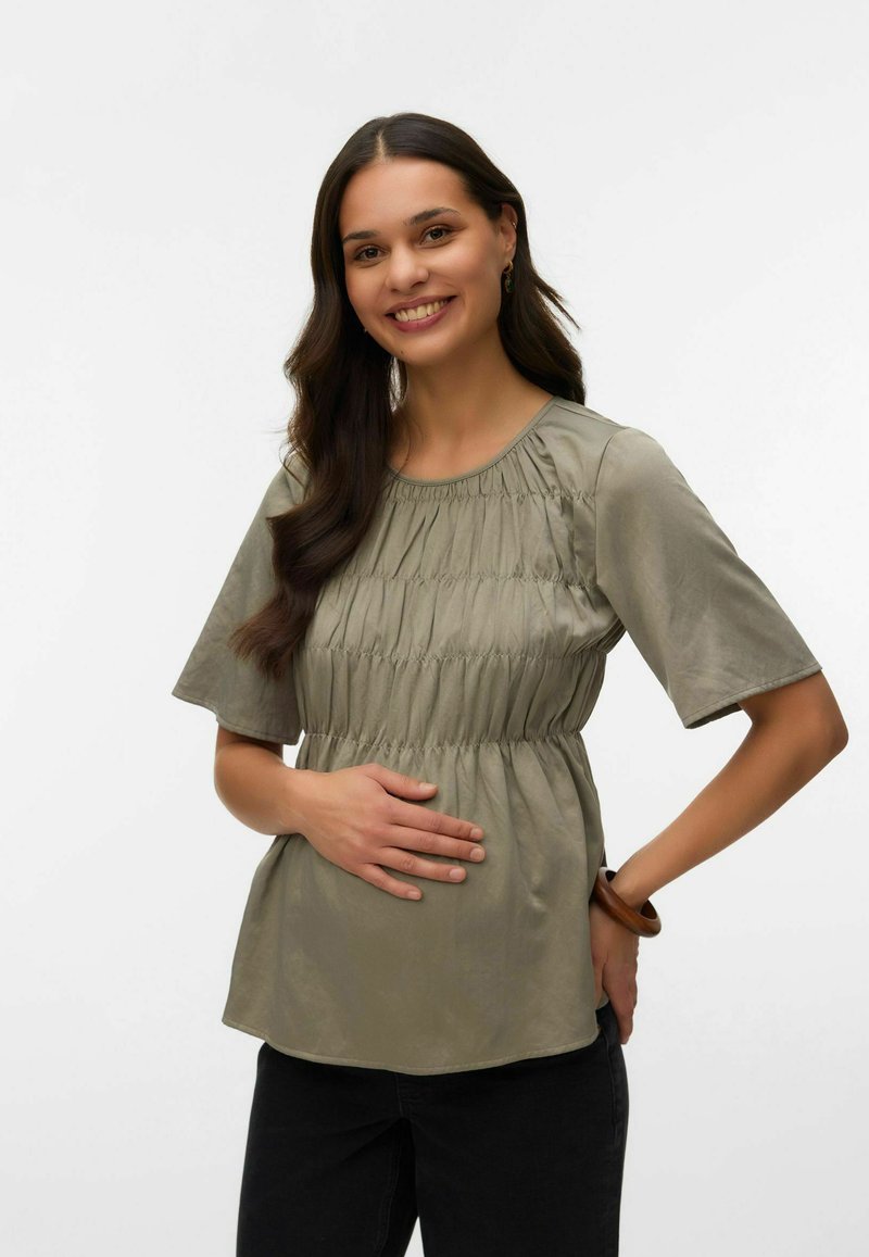 Naeratav naine pikkade tumedate juustega kannab kogutud salvei rohelise tooni pluusi ja musti pükse, üks käsi kõhul ja teine puusal.