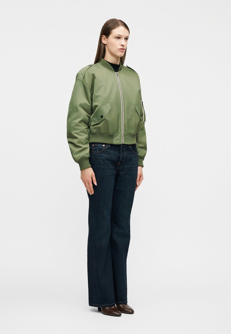 Femme debout portant une veste bomber vert clair, un jean bleu foncé et des chaussures à talons marron, sur un fond blanc uni.