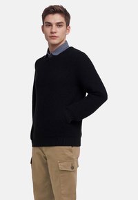 Black knitted sweater with a textured pattern, ribbed cuffs, and hem. Paired with light-colored cargo pants. Classic crew neck design.