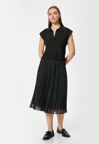 Schwarzes gestricktes Oberteil mit Kragen, kombiniert mit einem schwarzen, plissierten Rock mit weißen Punkten. Das Modell trägt schwarze Slipper.