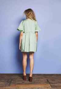 Ženska s curly lasmi stoji ob svetlobno modri steni, oblečena v kratko svetlo zeleno obleko in rjave platform sandale na leseni podlagi.