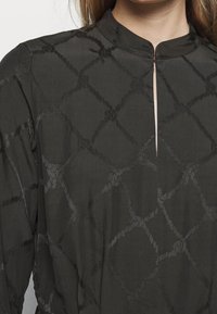 Blusa de tela texturizada oscura con un cuello alto, que presenta un sutil patrón de diamantes, una abertura frontal y un drapeado suave.