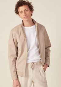 Beige leichte Jacke mit einem Reißverschlusskragen, kombiniert mit einem weißen T-Shirt und beigen Jogginghosen mit Kordelzug. Glatte Textur mit minimalen Details.