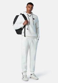 Felpa bianca con coulisse nere e logo, abbinata a pantaloni jogger bianchi con tasche e sneakers color crema. Zaino nero indossato su una spalla.