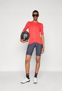 Rød sykkeltrøye med korte ermer, blå shorts og svarte sykkelsko. Modellen holder en svart hjelm og har på seg mørke solbriller.