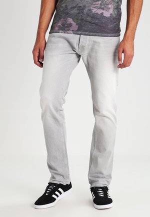 Homme portant un jean slim gris clair, des baskets noires et blanches, et une chemise sombre à motifs floraux, se tenant devant un fond uni.