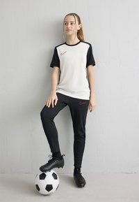 Tinejdžerica u crno-bijeloj sportskoj odjeći stoji naslonjena na zid s jednom nogom na nogometnoj lopti, gledajući u stranu.