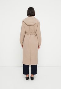 Trench-coat beige avec une capuche, une taille ceinturée et des manches longues. Il présente une coupe droite et une fente à l'arrière. Porté avec un pantalon bleu marine.
