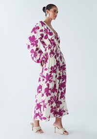Robe maxi crème avec de grands motifs floraux violets, présentant un décolleté en V profond, des manches longues et une taille smockée. Portée avec des talons légers.