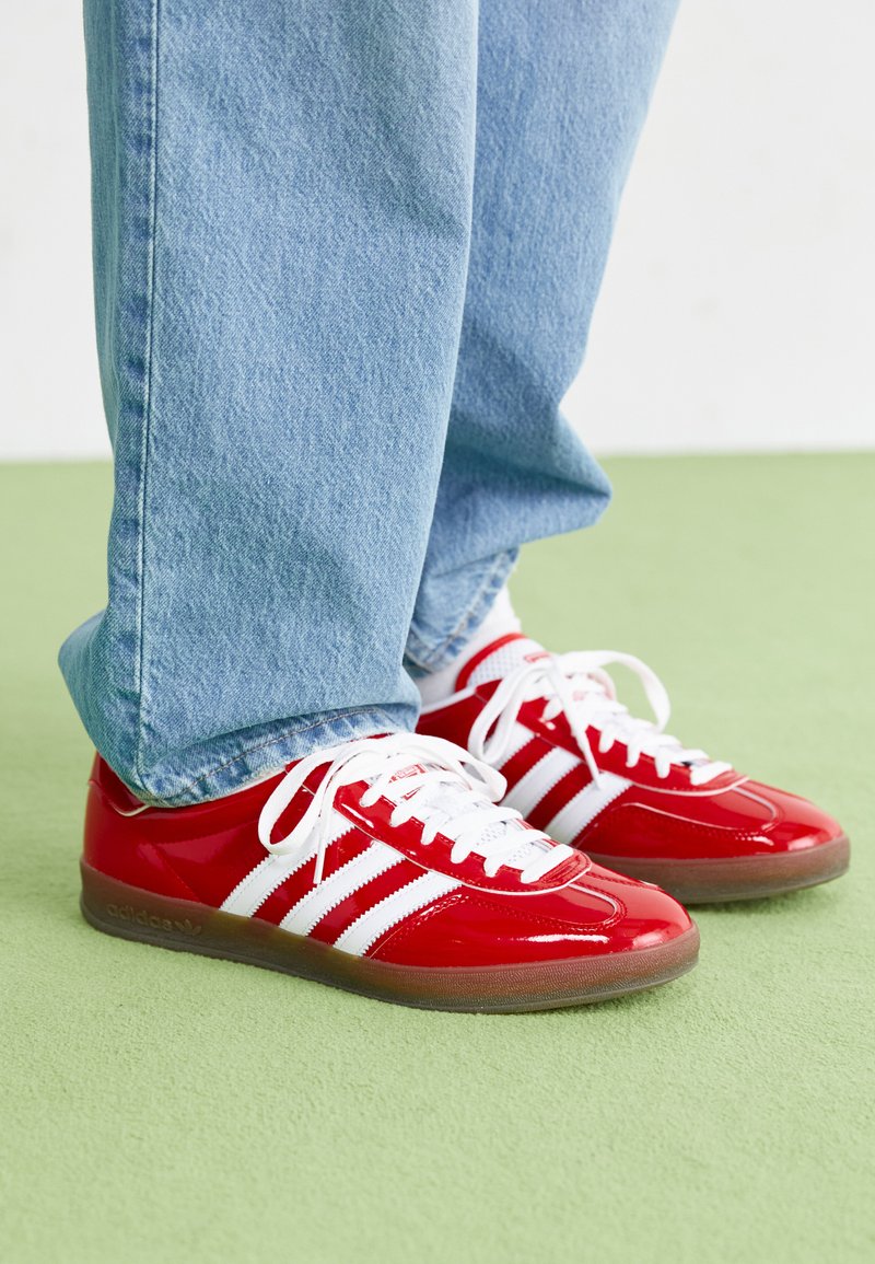 Red patent leather sneakers with white stripes, white laces, and a textured gray rubber sole, paired with light blue denim pants.