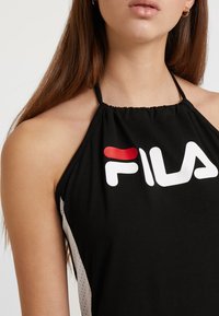 Czarny top bez rękawów z halter neck, z dużym, białym i czerwonym logo FILA oraz teksturowanymi akcentami siateczkowymi po bokach.