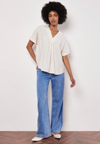 Blusa crema rilassata con scollo a V e dettaglio plissettato, abbinata a jeans blu a gamba larga e décolleté bianchi a punta. Sfondo in legno.