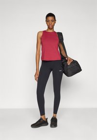 Röd atletisk linne, svarta leggings med logo och svarta sneakers. Modellen håller en svart duffelväska. Enkel, figurnära design med släta texturer.