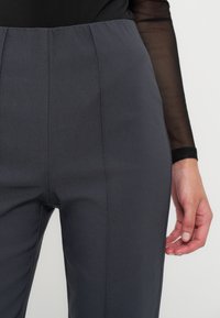 Personne portant un pantalon gris foncé taille haute et un haut noir transparent à manches longues, visible de la taille à la cuisse avec le bras droit visible.