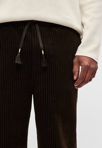 Brūnas kordurīna bikses ar vertikālām ribām, kurām ir auklas jostasvieta ar bārkstīm. Jostas aukliņām ir dzelteni akcenti.