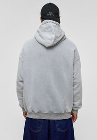 Lichtgrijze hoodie met een relaxte pasvorm, ribbels bij de mouwen en onderkant. De achterkant heeft geen afbeeldingen, terwijl een zwarte pet met witte tekst wordt gedragen.