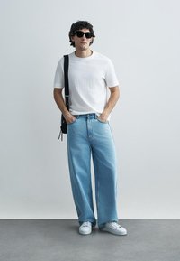 T-shirt branca texturizada combinada com jeans azul claro de perna larga, sapatilhas brancas e óculos de sol pretos. Mala preta de ombro realça o visual.