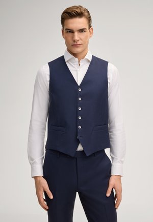 Giovane uomo con camicia bianca e gilet blu scuro abbottonato con pantaloni blu scuro coordinati, in piedi su uno sfondo chiaro e semplice.
