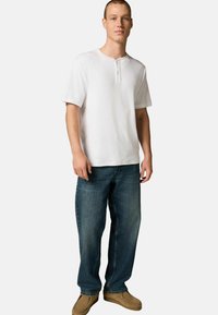Camisa henley blanca de manga corta, jeans de mezclilla azul y zapatos de slip-on marrón claro. La camisa tiene un cuello con una abertura de dos botones.