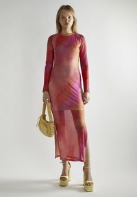 Sukienka o długim rękawie, dopasowana, w kolorze różowo-pomarańczowym w stylu tie-dye, z bocznym rozcięciem. Modelka trzyma plecioną żółtą torebkę oraz pasujące, masywne obcasy.