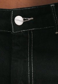 Jeans di denim neri con cuciture bianche, dotati di chiusura a pulsante in vita. Il tessuto sembra robusto e ha una consistenza liscia.