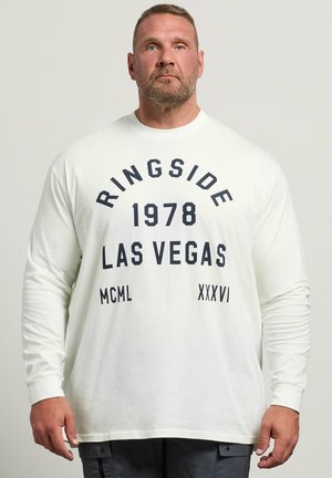 Hombre con pelo corto y barba que lleva una camisa blanca de manga larga con el texto «"Ringside 1978 Las Vegas MCML XXXVI"», de pie frente a un fondo sencillo.
