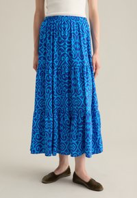 Gonna blu con motivo a fantasia, dotata di una vita elastica, lunga fino al pavimento, con un design a strati e intricati motivi geometrici. Abbinata a scarpe scure senza lacci.